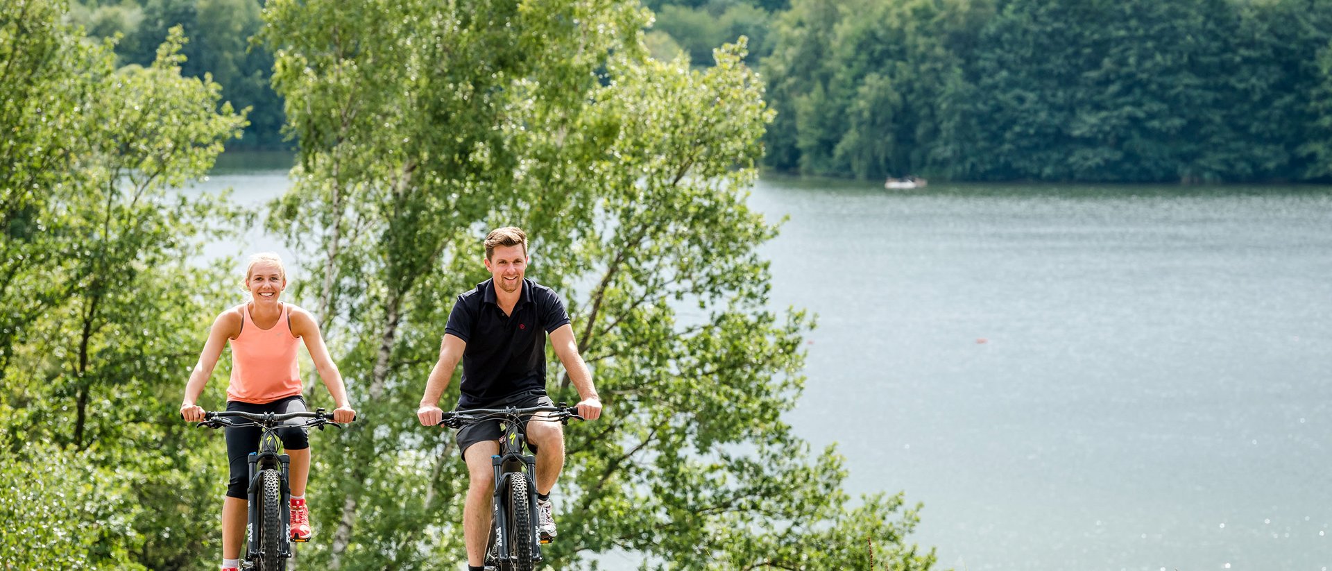 Zwei Personen fahren mit Fahrrädern auf einem Weg neben einem See und Bäumen