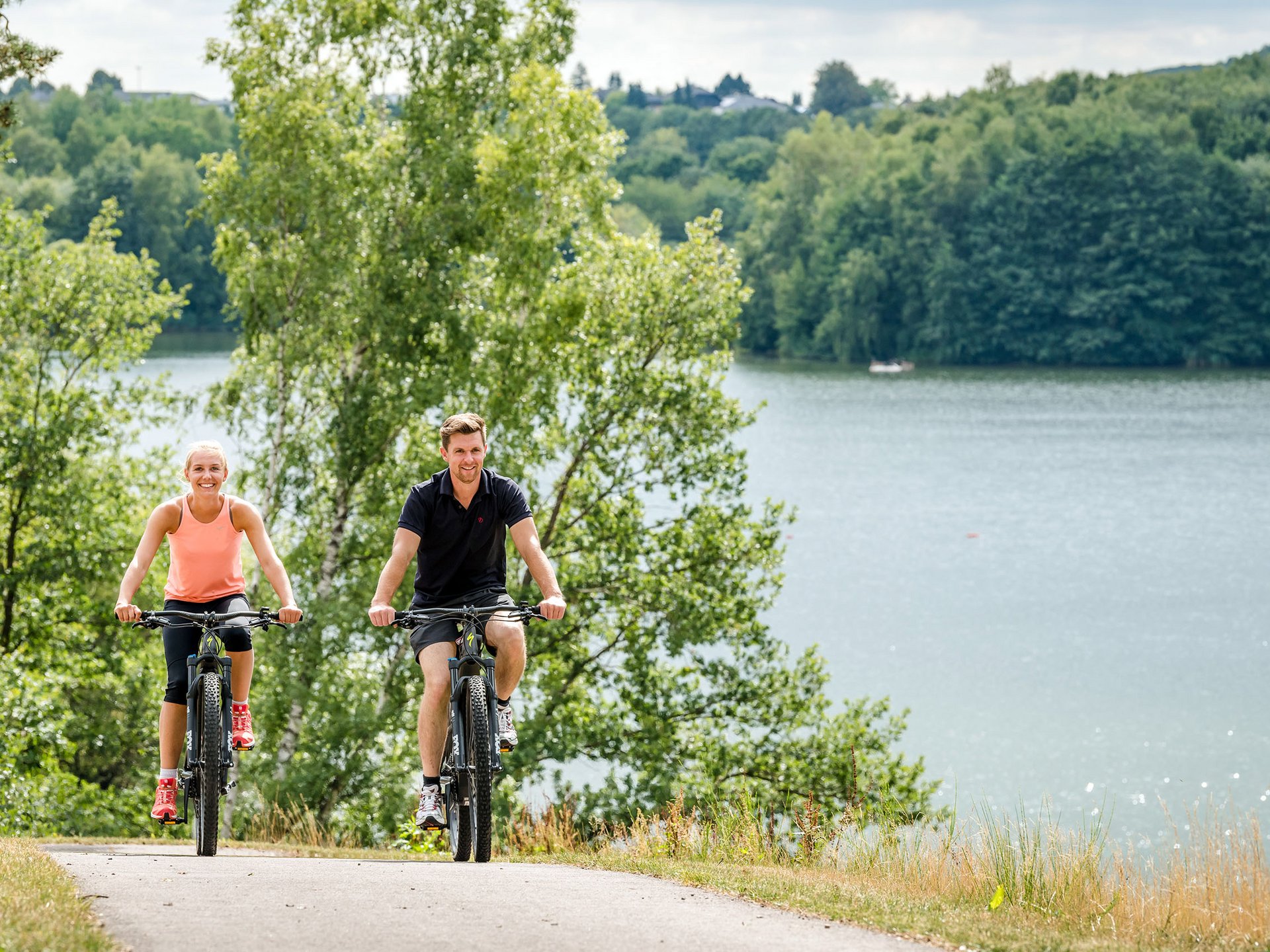 Two people cycling on a path beside a lake with trees in the background