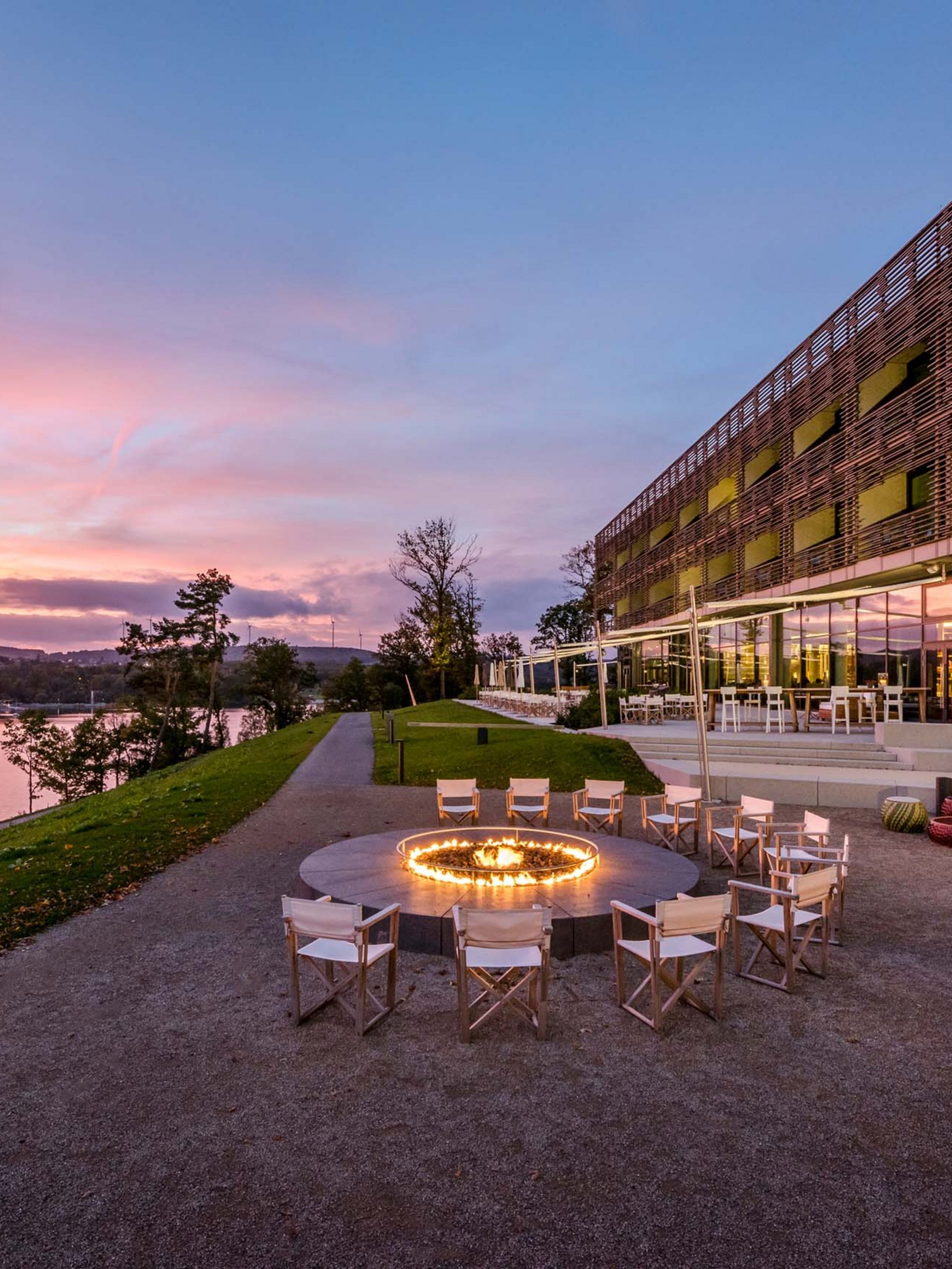 Seating area with fire pit beside modern building and river at sunset