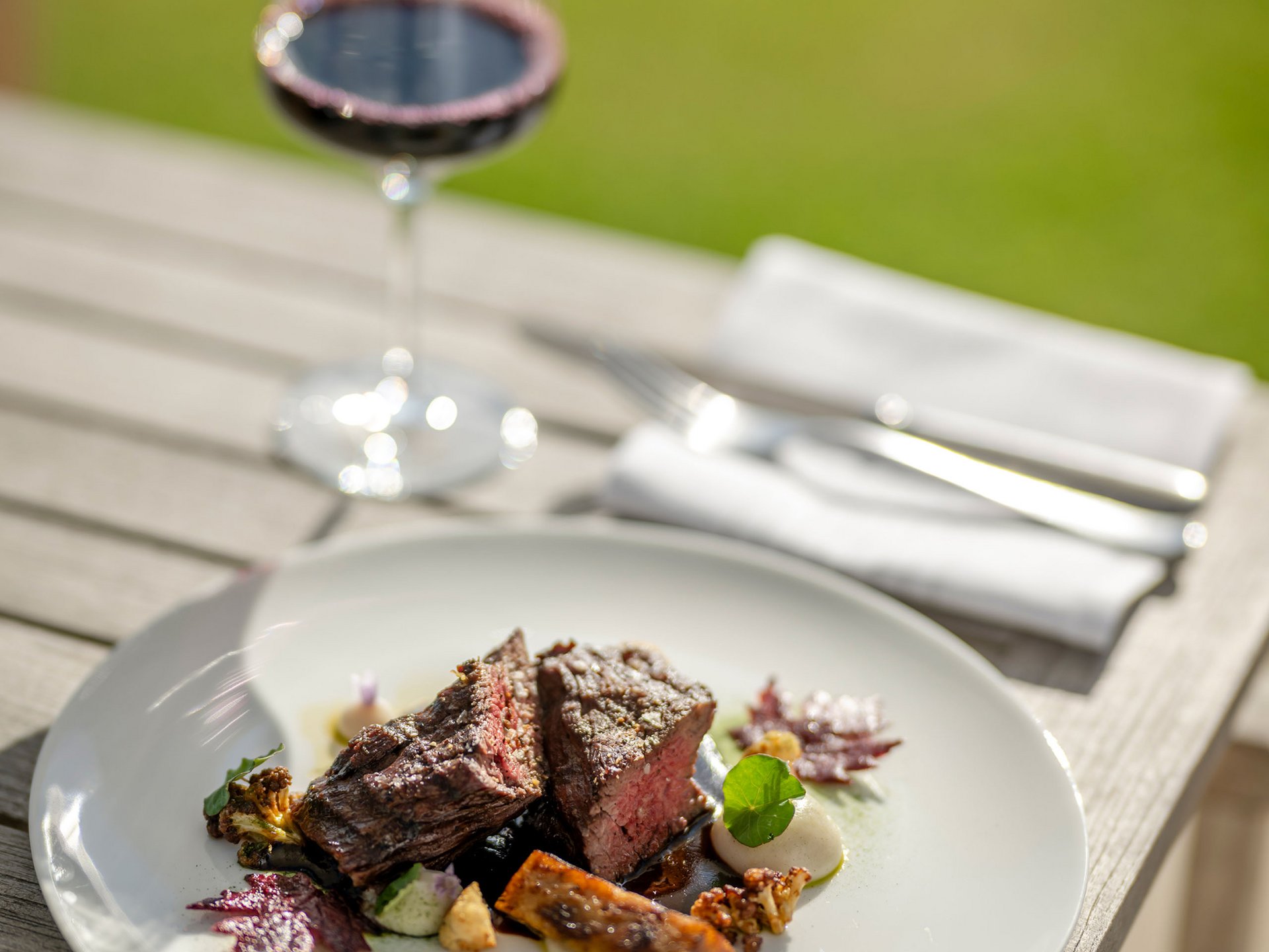 Steak with vegetables on white plate and glass of red wine outdoors