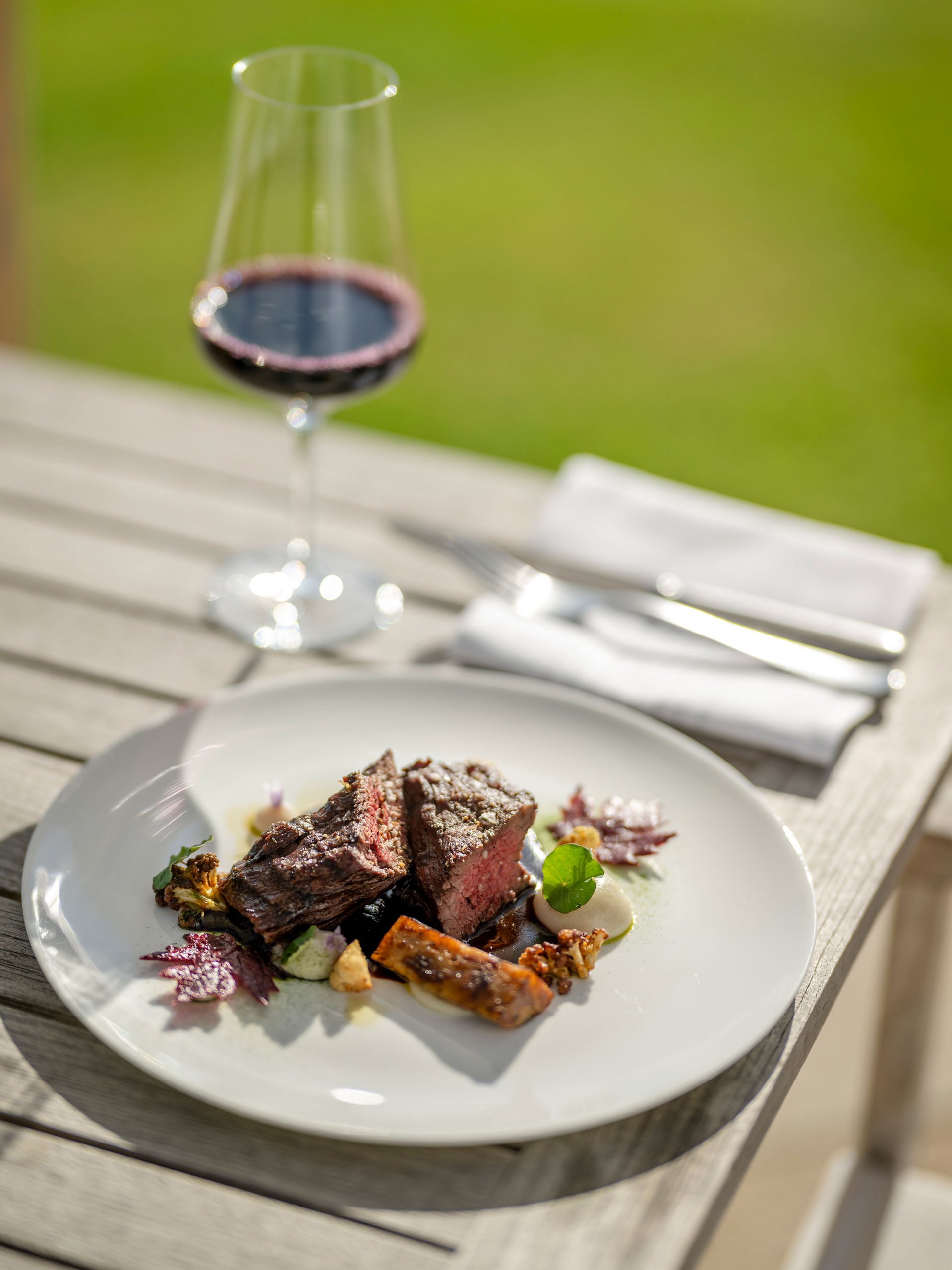 Steak with vegetables on white plate and glass of red wine outdoors