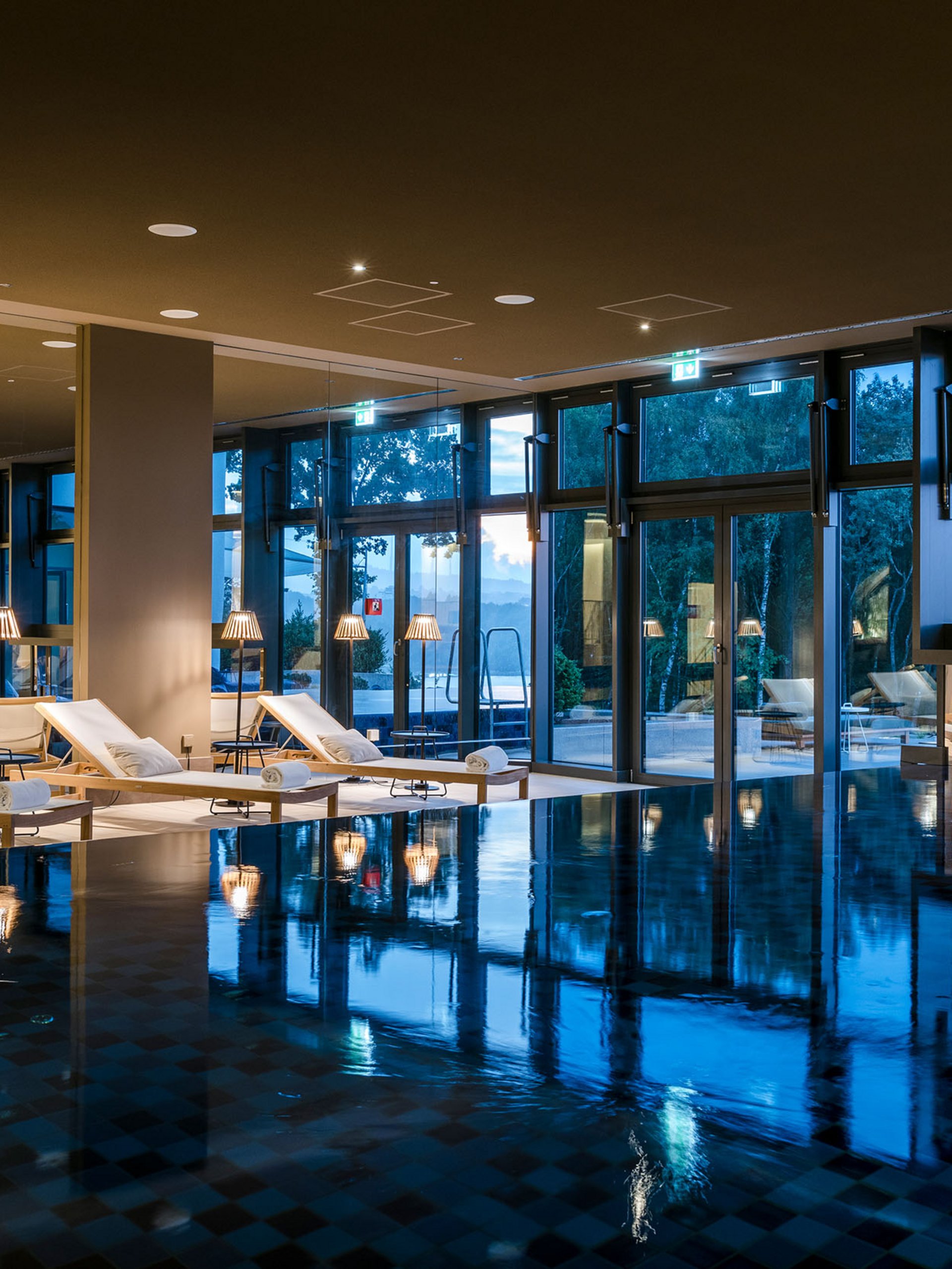 Indoor pool with lounge chairs and large windows at dusk