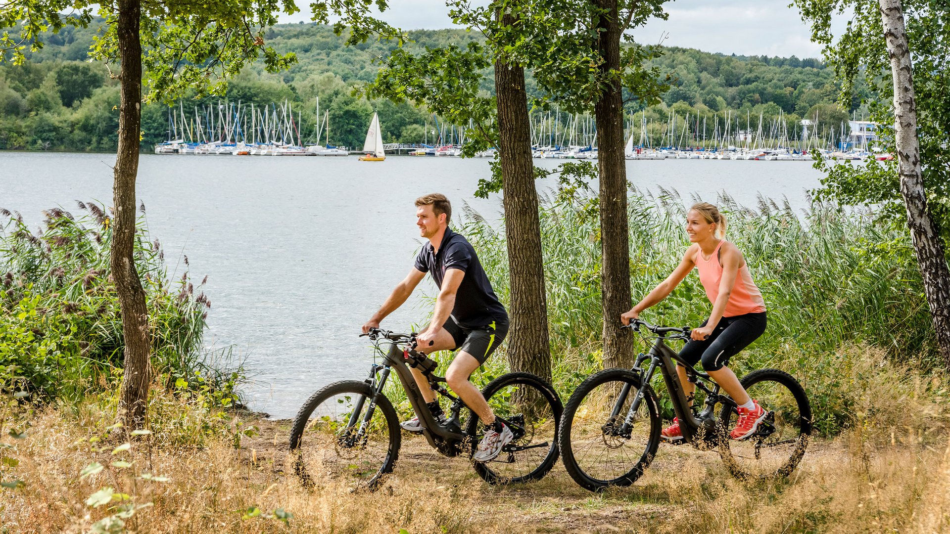 Couple riding mountain bikes by lake with sailboats in the background