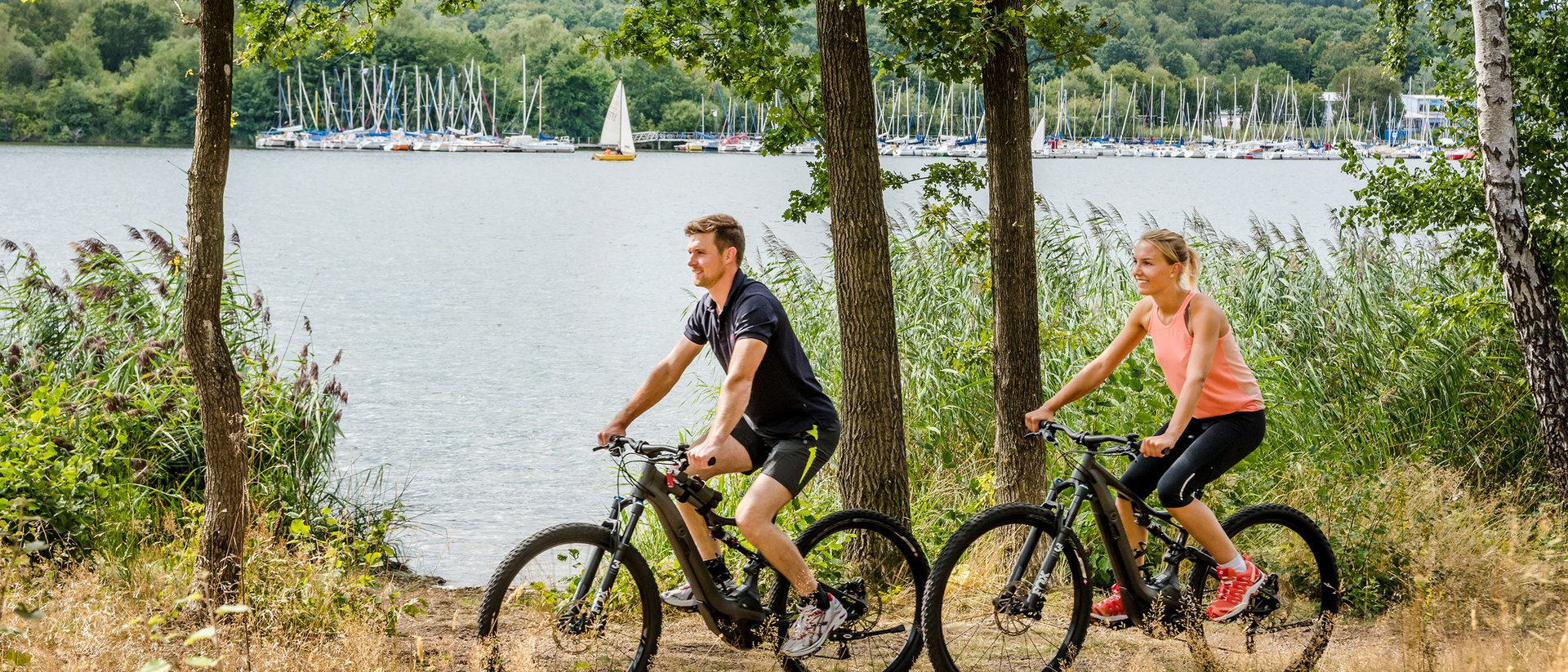 Paar fährt Mountainbikes am Seeufer mit Segelbooten im Hintergrund