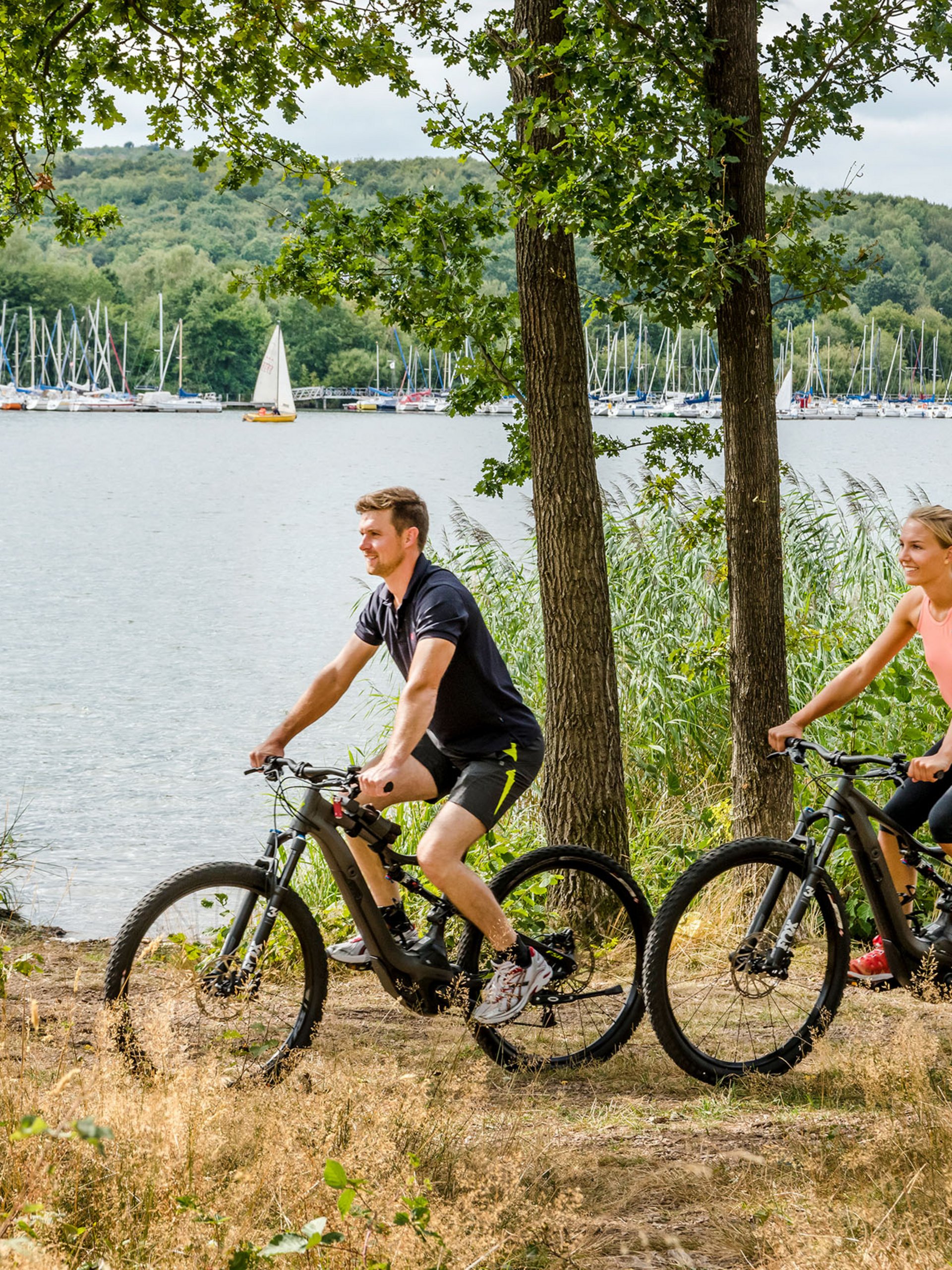 Couple riding mountain bikes by lake with sailboats in the background