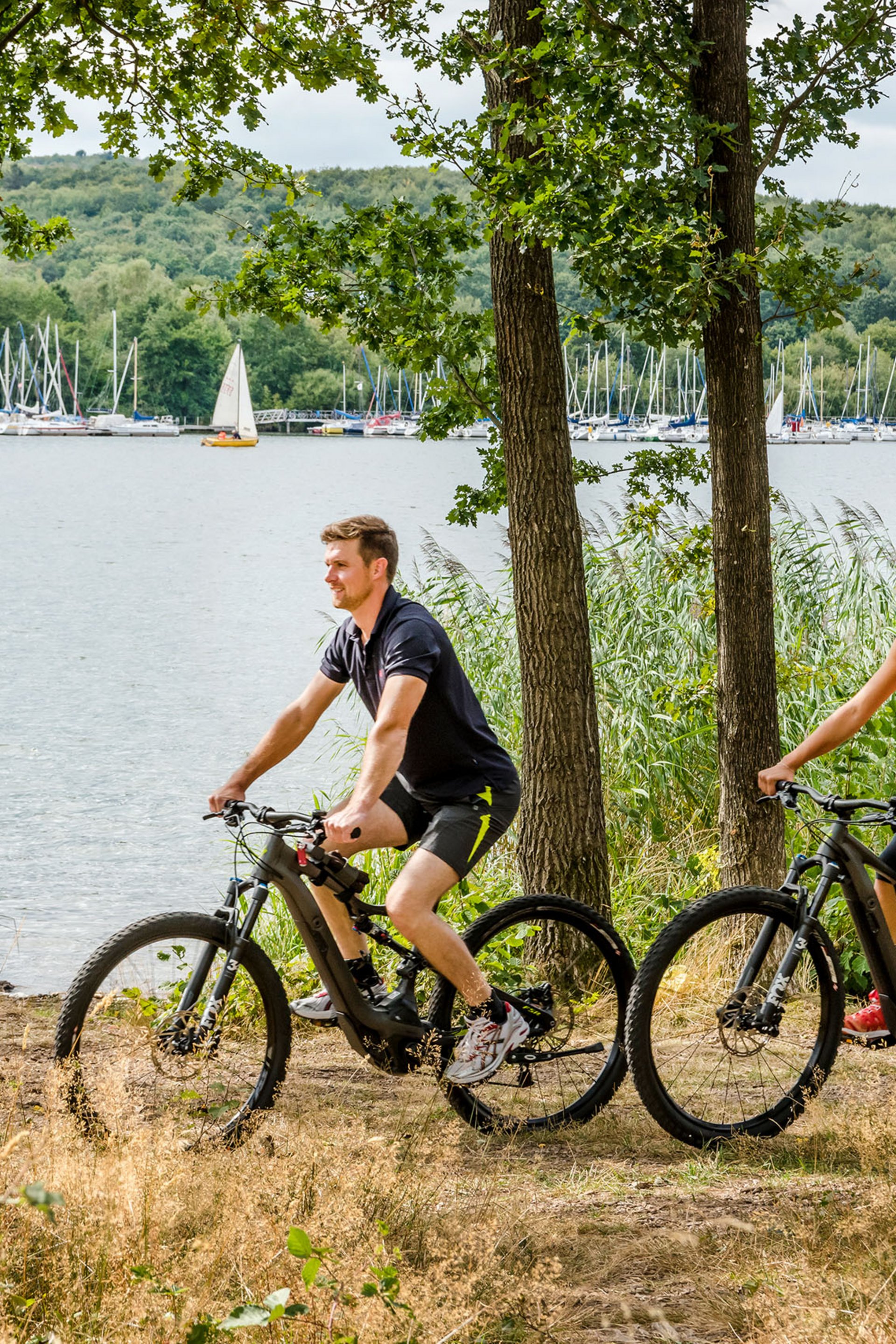 Paar fährt Mountainbikes am Seeufer mit Segelbooten im Hintergrund