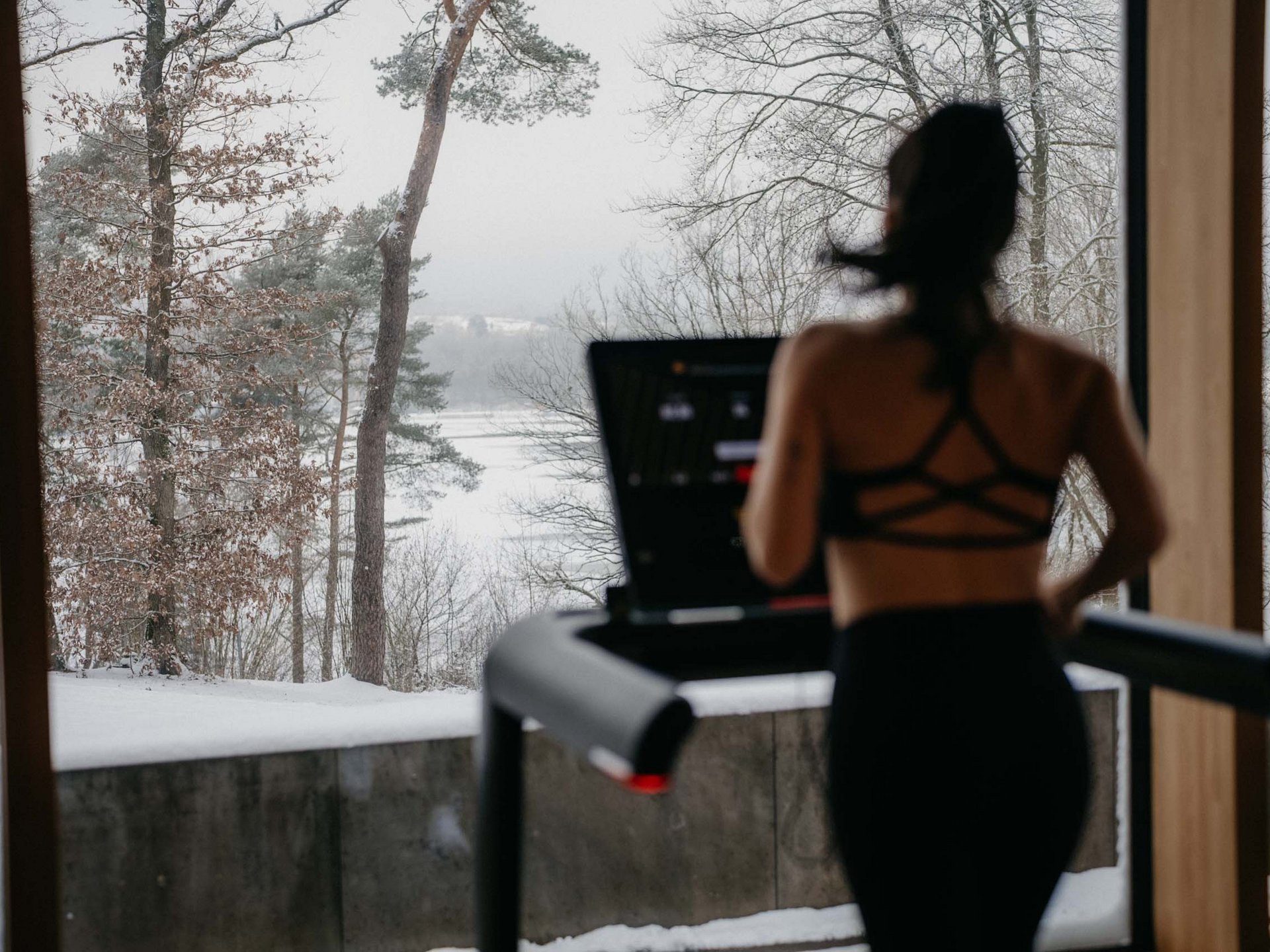 Seezeit re.connect – Bewusste Verbindung mit Natur & Kraftort Frau trainiert auf einem Laufband vor einem Fenster mit verschneiter Waldlandschaft