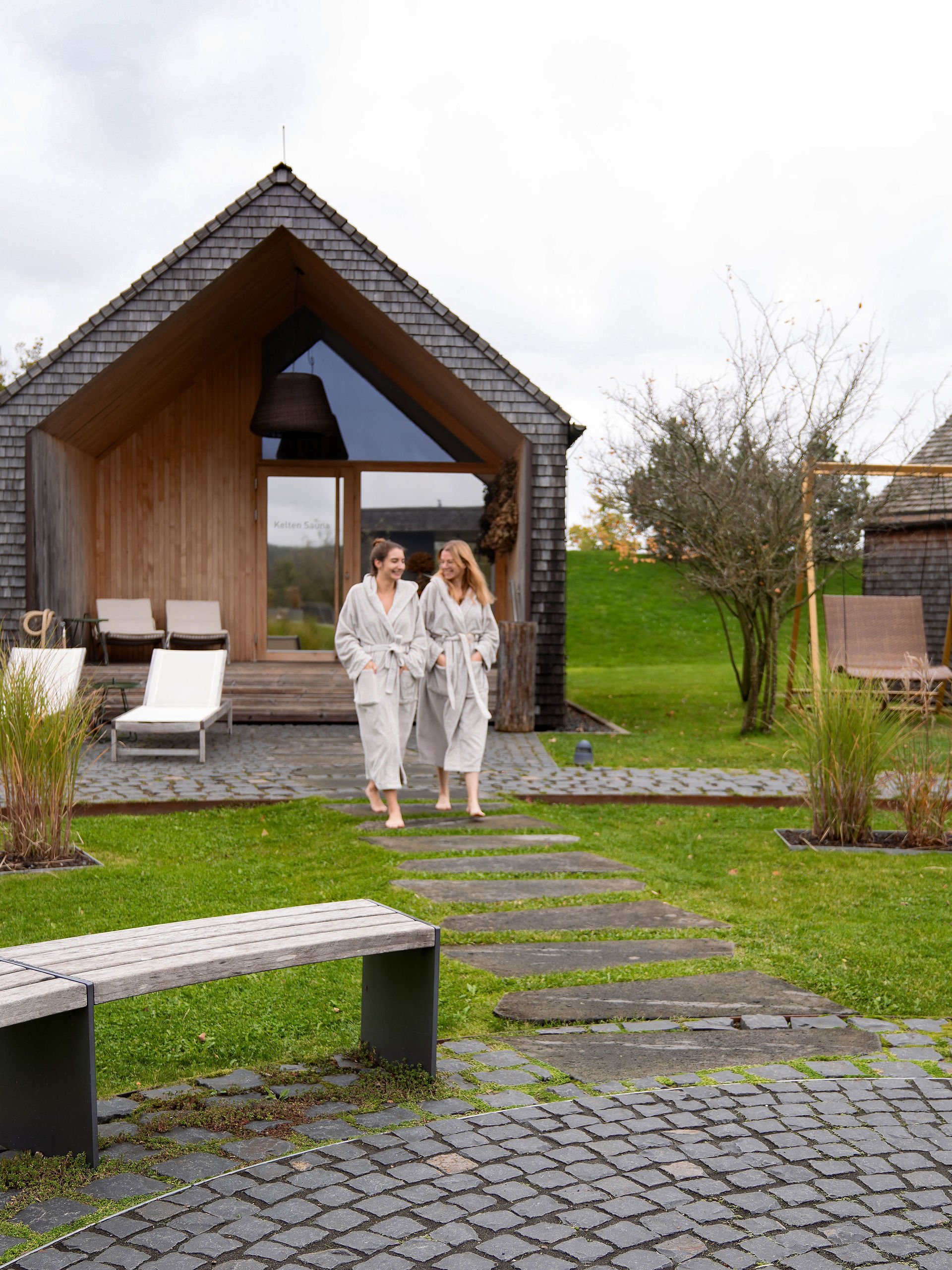 Two women in bathrobes outside a modern sauna surrounded by greenery