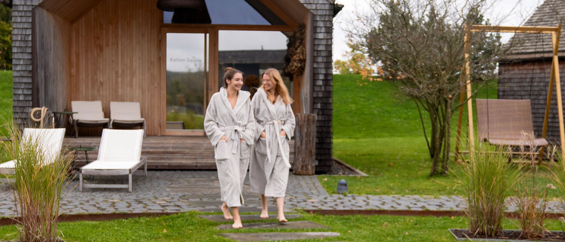 Two women in bathrobes outside a modern sauna surrounded by greenery