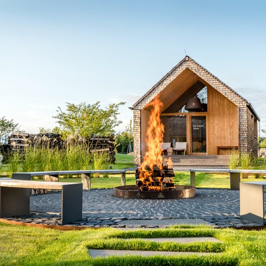 Offene Feuerstelle mit hölzernen Bänken vor einer kleinen Holzsauna im Garten