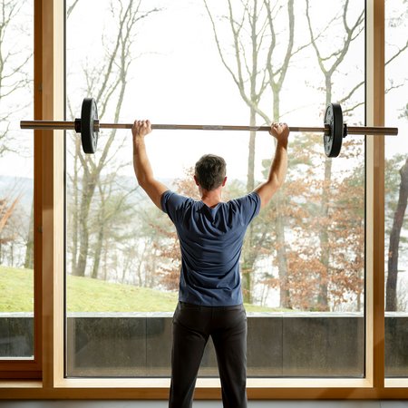 Mann hebt Langhantel vor großem Fenster mit Naturblick