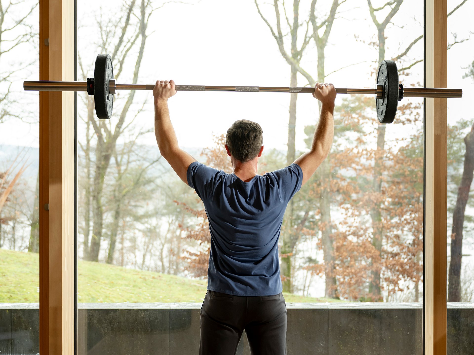 Mann hebt Langhantel vor großem Fenster mit Naturblick
