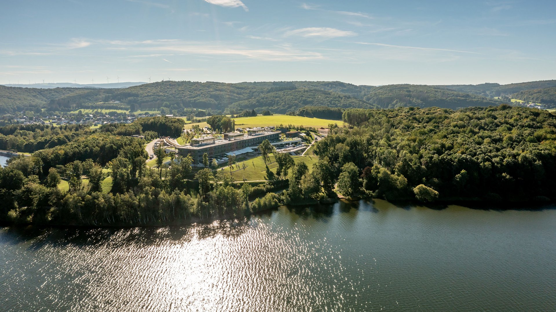 Luftaufnahme eines Sees mit bewaldeten Ufern und einem großen Gebäude bei Sonnenschein