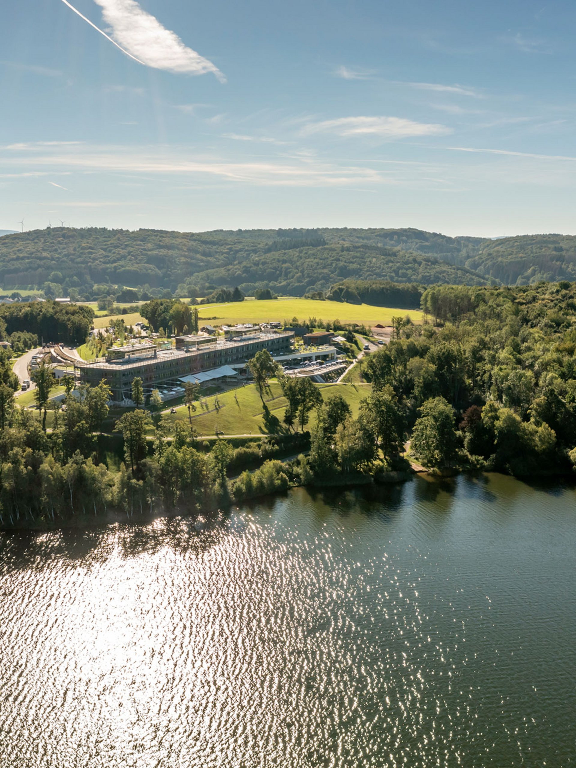 Luftaufnahme eines Sees mit bewaldeten Ufern und einem großen Gebäude bei Sonnenschein