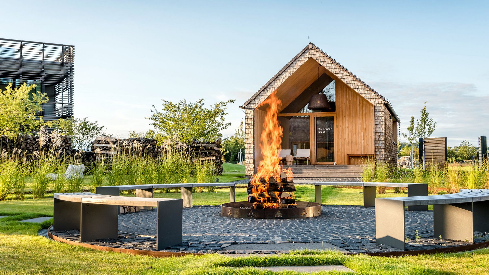 Fire pit with burning wood and circular bench in front of a wooden sauna