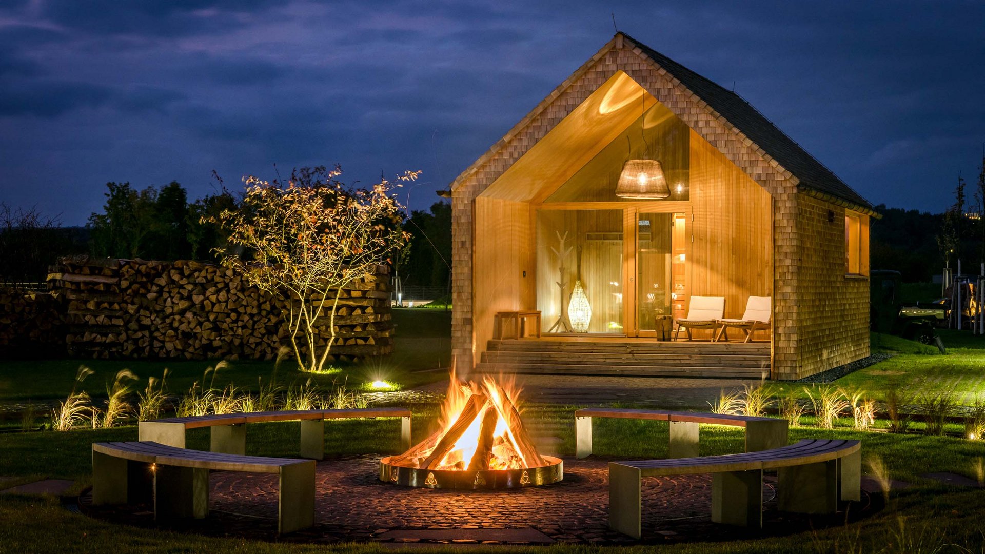 Wooden cabin at night with campfire and benches in the garden