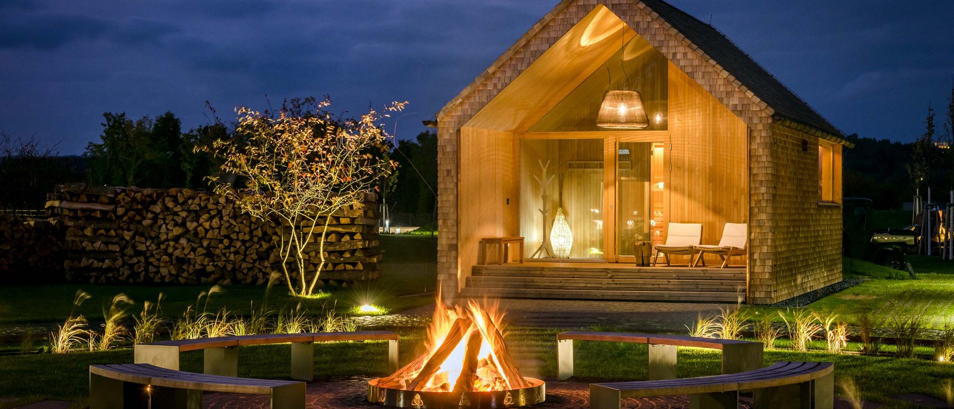 Holzhaus bei Nacht mit Lagerfeuer und Sitzbänken im Garten
