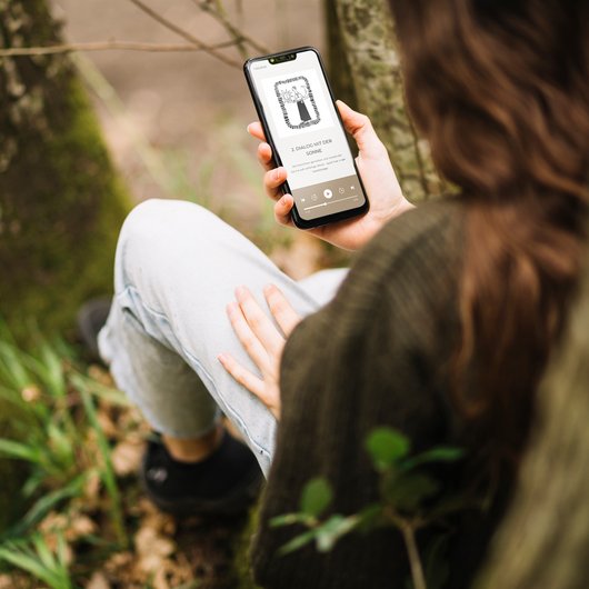 Audiowalk Person sitzt im Freien und hält ein Smartphone mit einer Audiowiedergabe