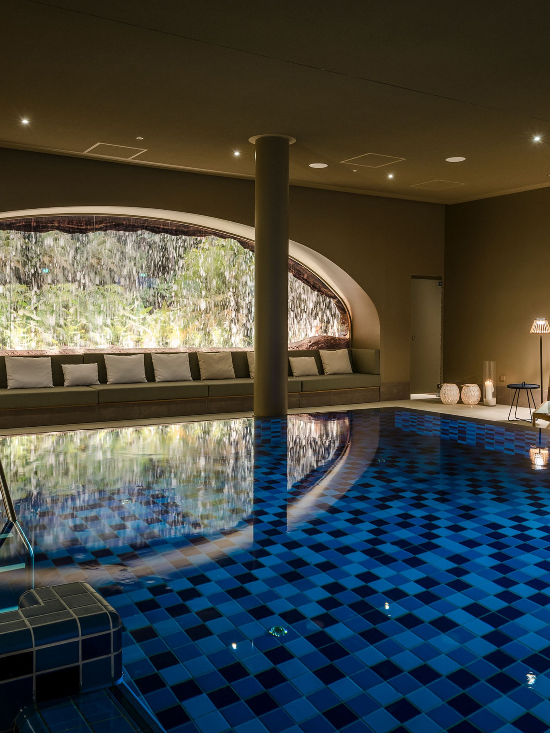 Heated indoor pool with waterfall and lounge chairs under warm lighting