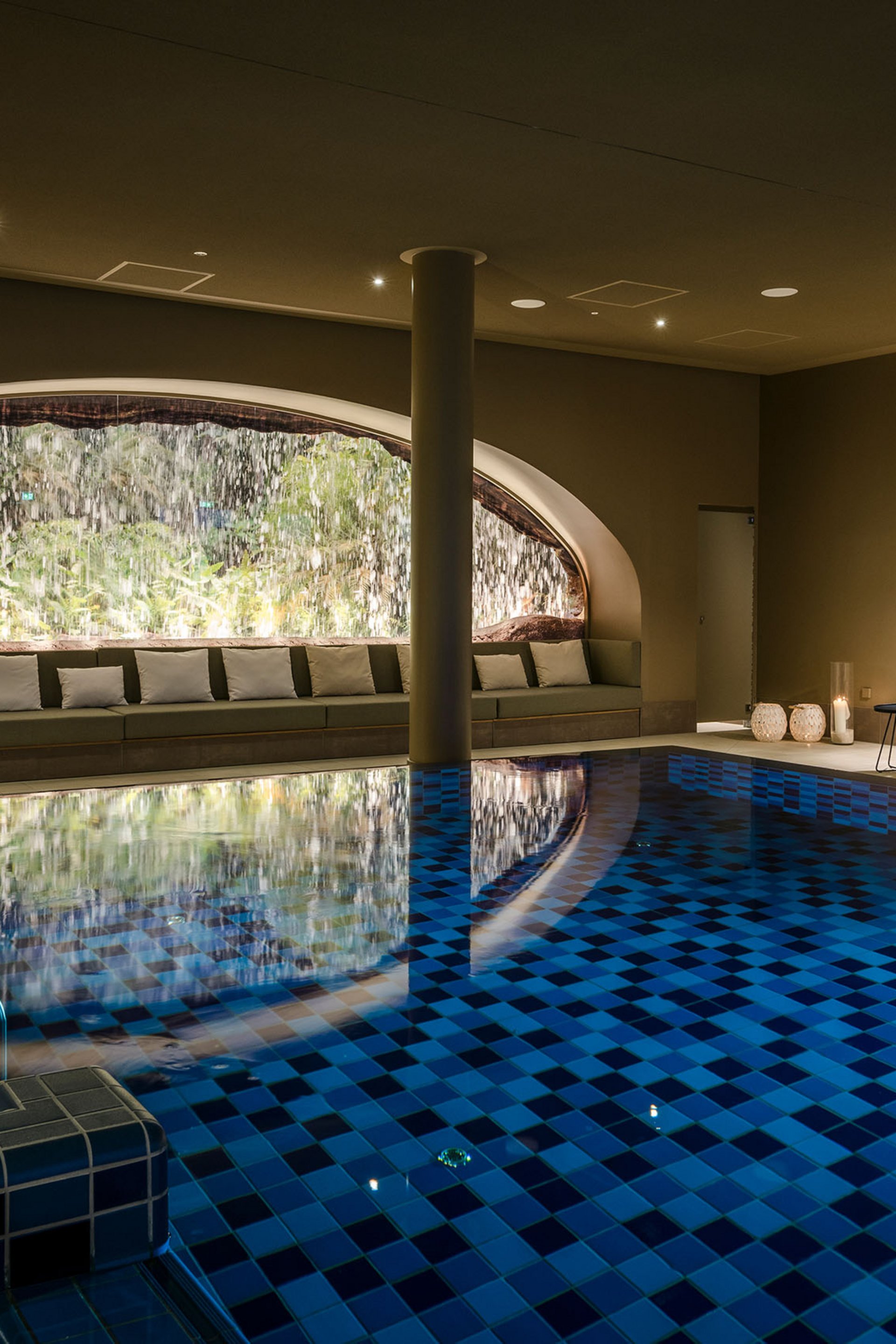 Heated indoor pool with waterfall and lounge chairs under warm lighting