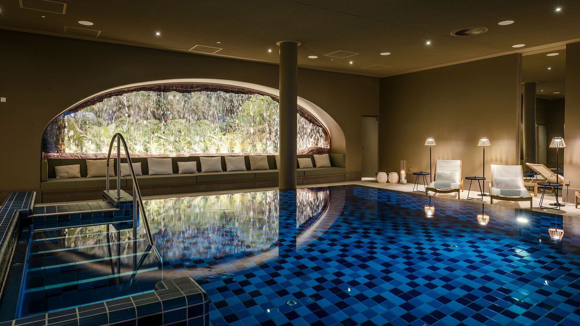 Heated indoor pool with waterfall and lounge chairs under warm lighting