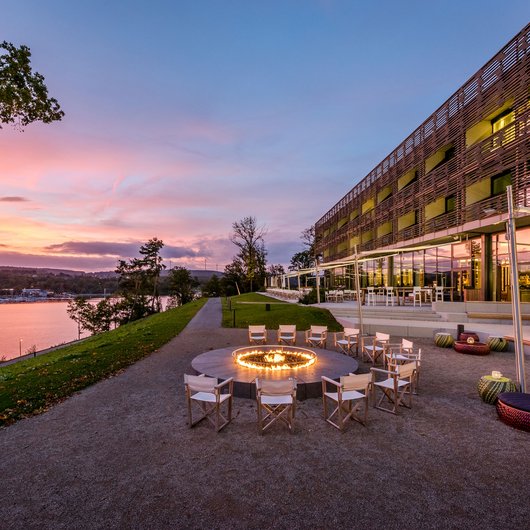 Feuerstelle mit Stühlen vor modernem Gebäude am Flussufer bei Sonnenuntergang