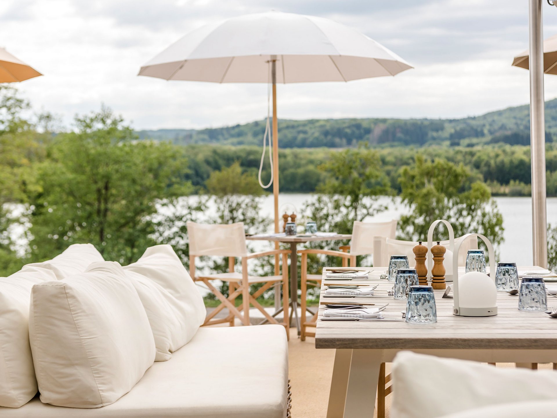Hell eingerichtete Terrasse mit Tisch und weißen Sitzkissen am See
