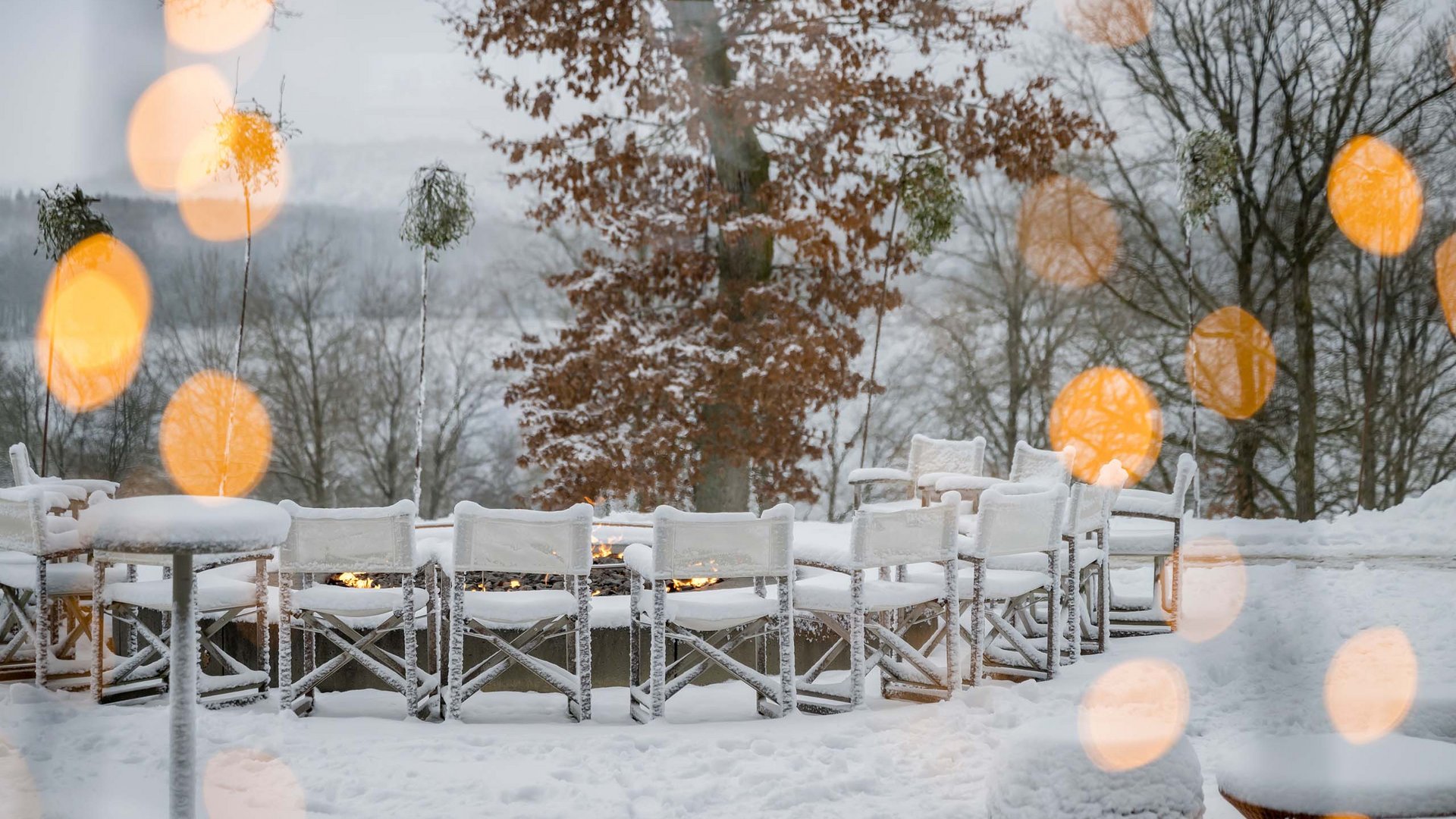 Verschneite Terrasse mit weißen Stühlen, Tischen und kleinen Feuern vor kahlen Bäumen