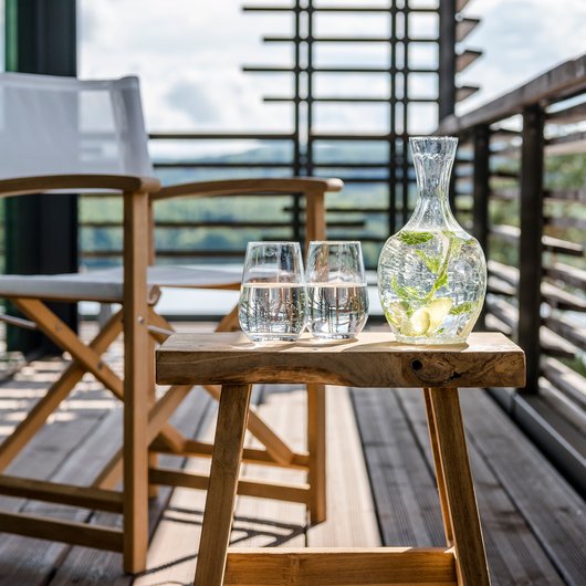 Une bouteille d'eau avec des tranches de concombre et deux verres sur un tabouret en bois sur un balcon.