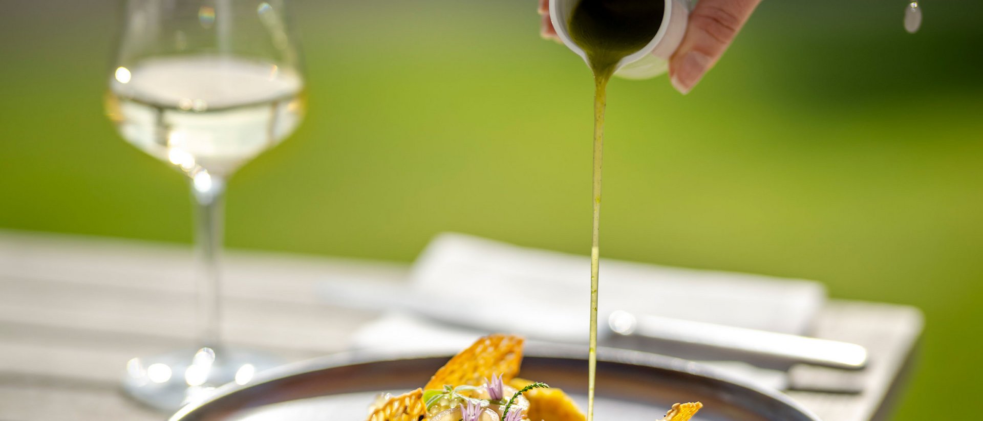 Hand pouring green sauce over an artfully plated gourmet dish outdoors