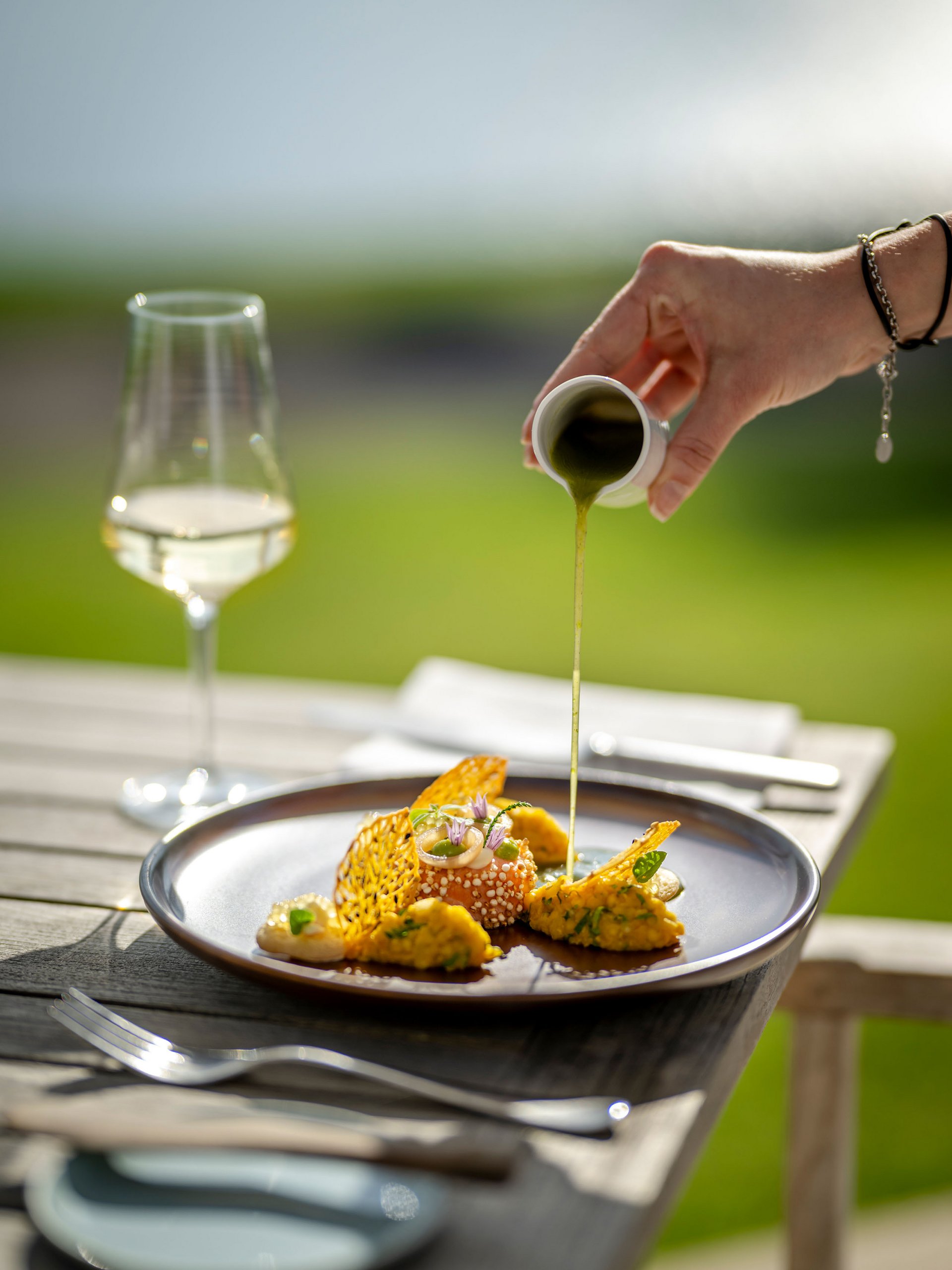 Hand pouring green sauce over an artfully plated gourmet dish outdoors