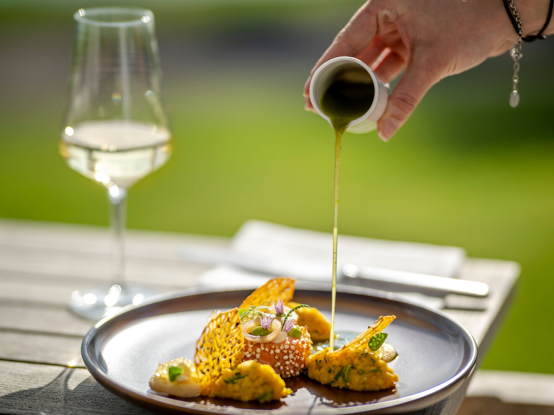 Hand pouring green sauce over an artfully plated gourmet dish outdoors
