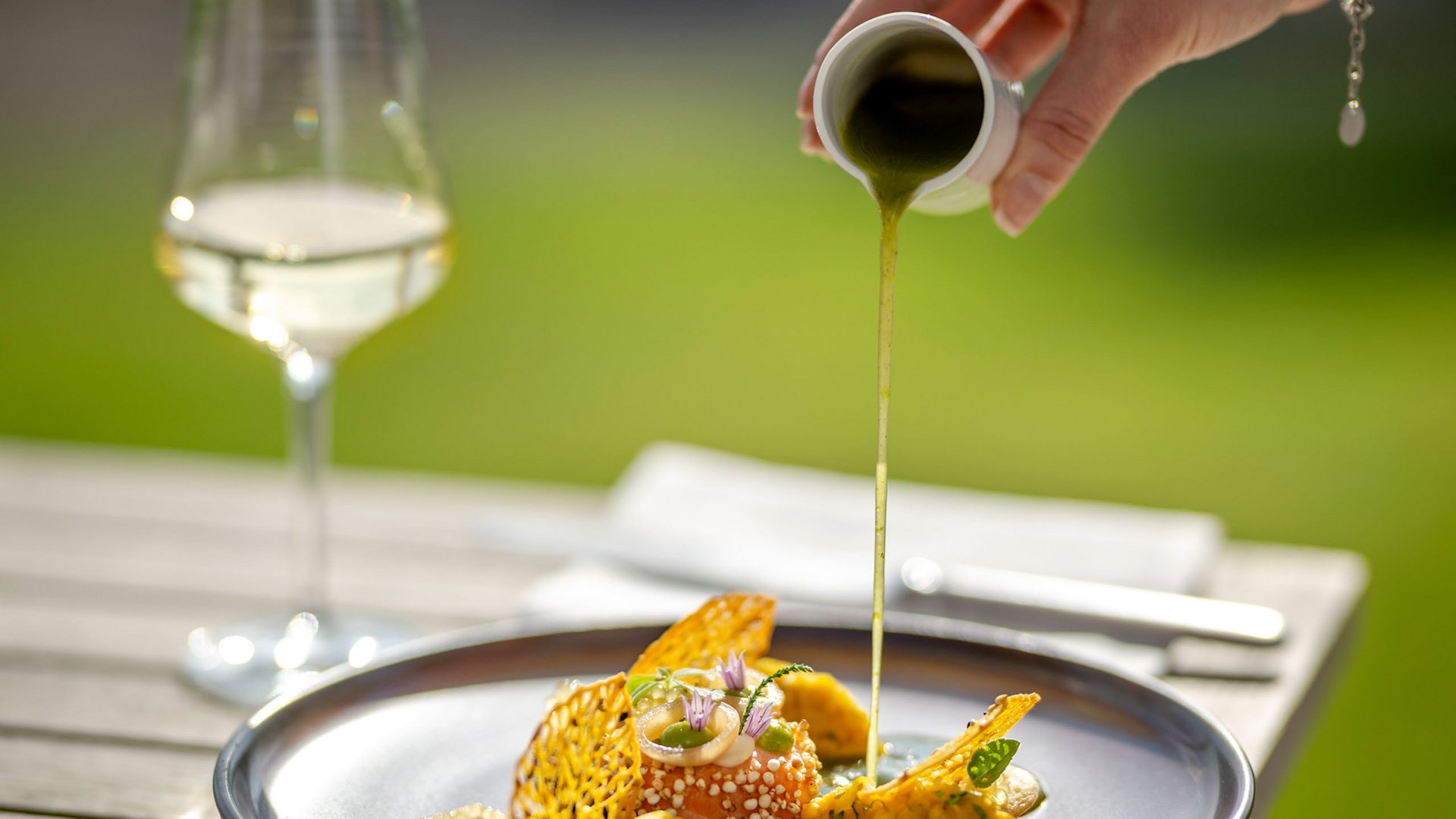 Hand gießt grüne Sauce über eine kunstvoll angerichtete Gourmet-Mahlzeit im Freien