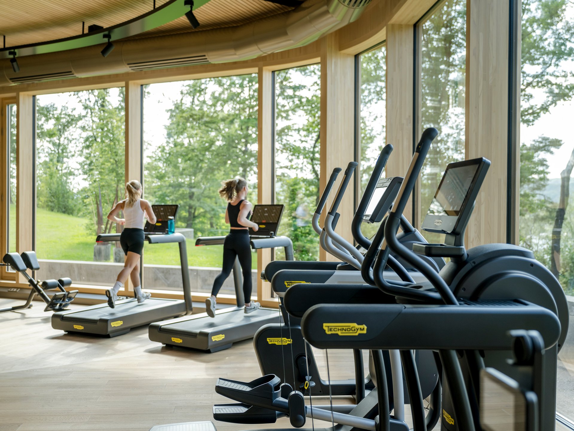 Zwei Frauen laufen auf Laufbändern in einem Fitnessstudio mit großen Fenstern