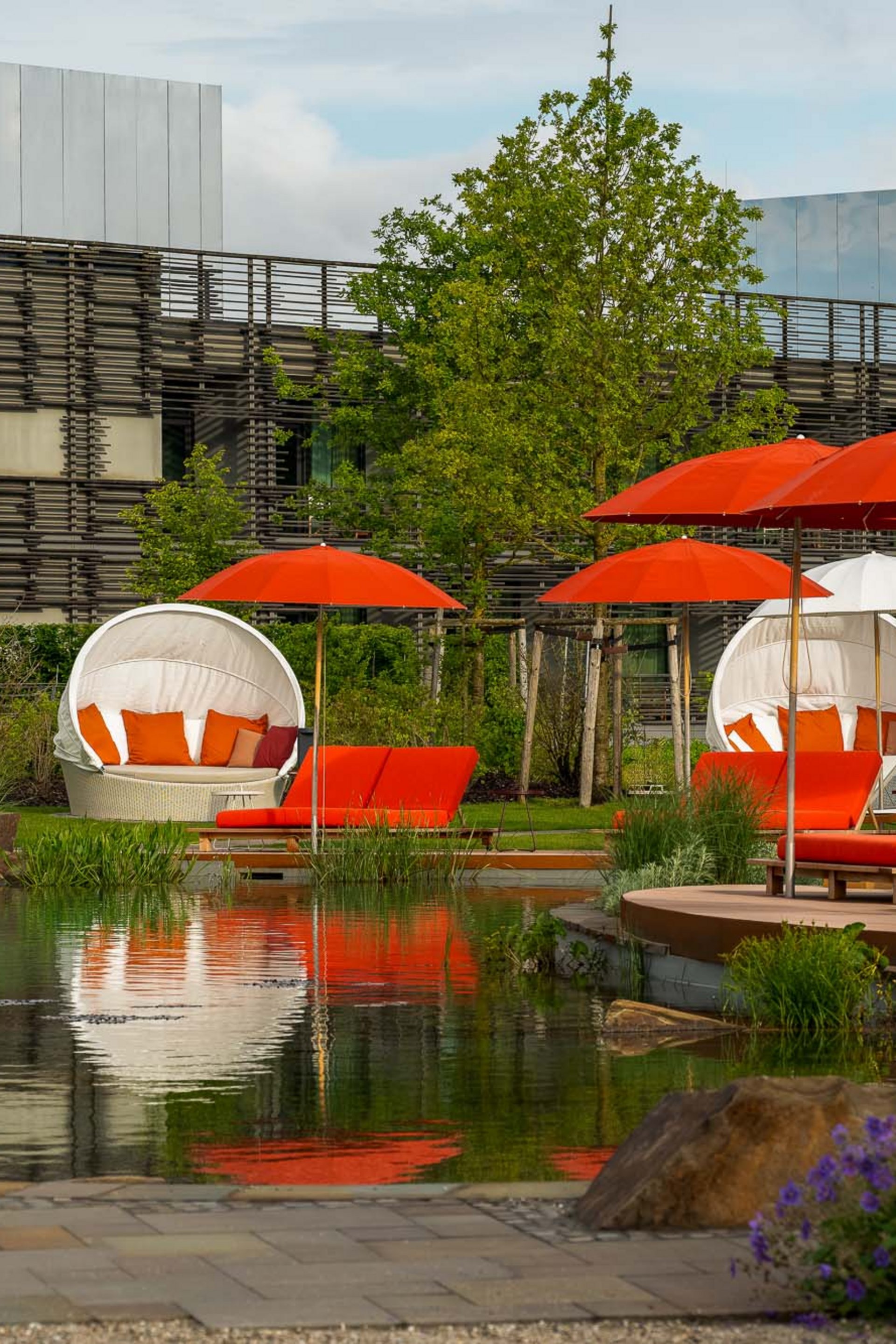 Red and white outdoor lounge chairs and umbrellas beside a pond in garden area