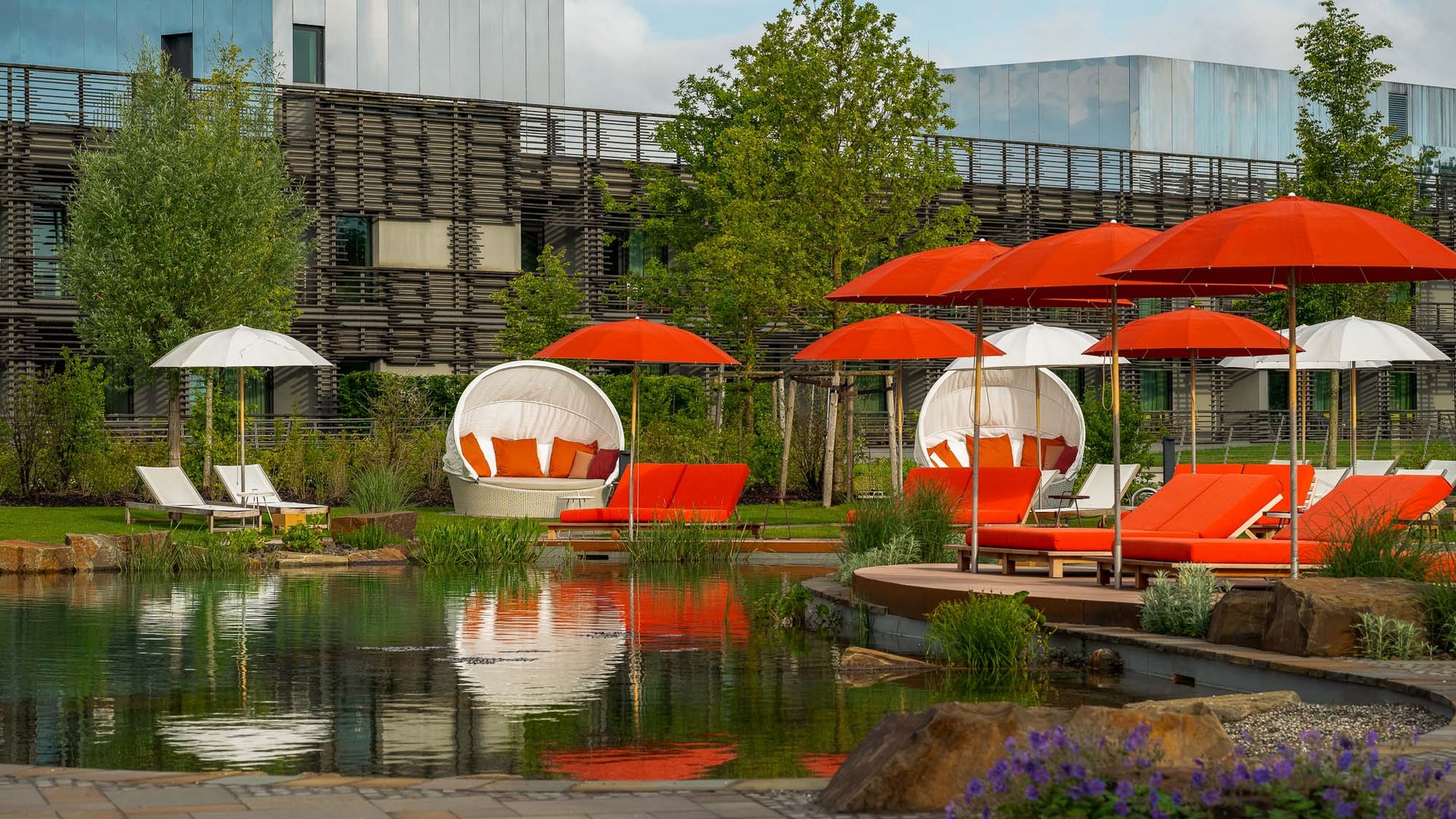 Red and white outdoor lounge chairs and umbrellas beside a pond in garden area