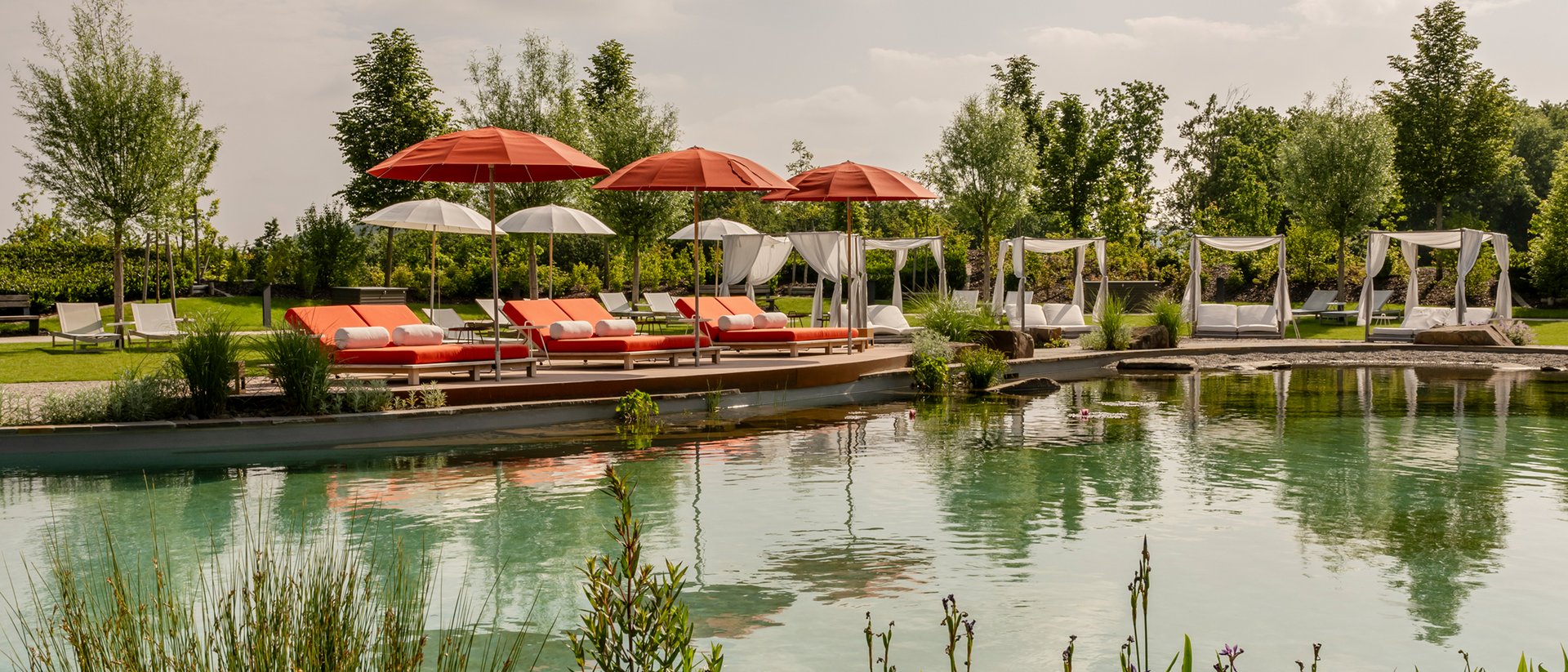 Ein natürlicher Pool mit roten Sonnenschirmen und Liegen im Garten bei bewölktem Himmel.