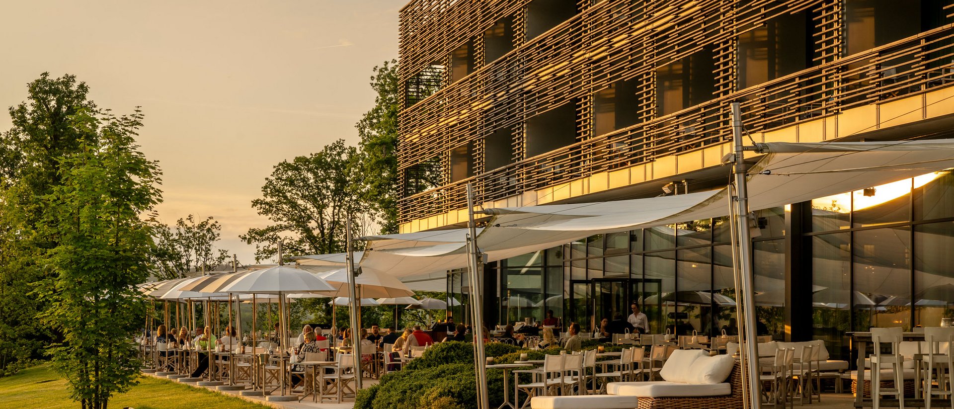 Sonnenterrasse eines Restaurants mit Gästen und moderner Holzfassade bei Sonnenuntergang