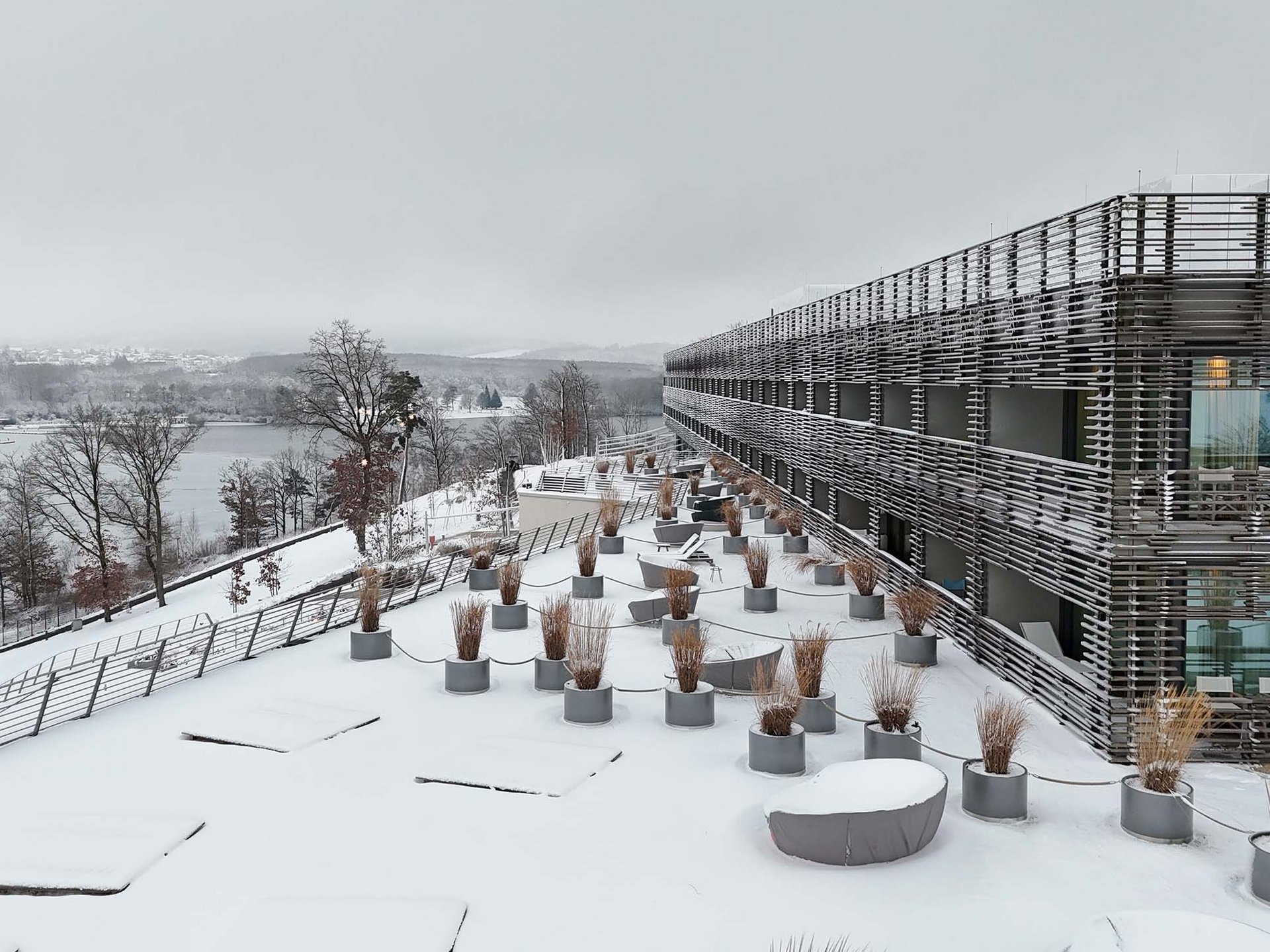 Verschneite Terrasse mit Pflanzkübeln neben einem modernen Gebäude und Blick auf eine winterliche Landschaft